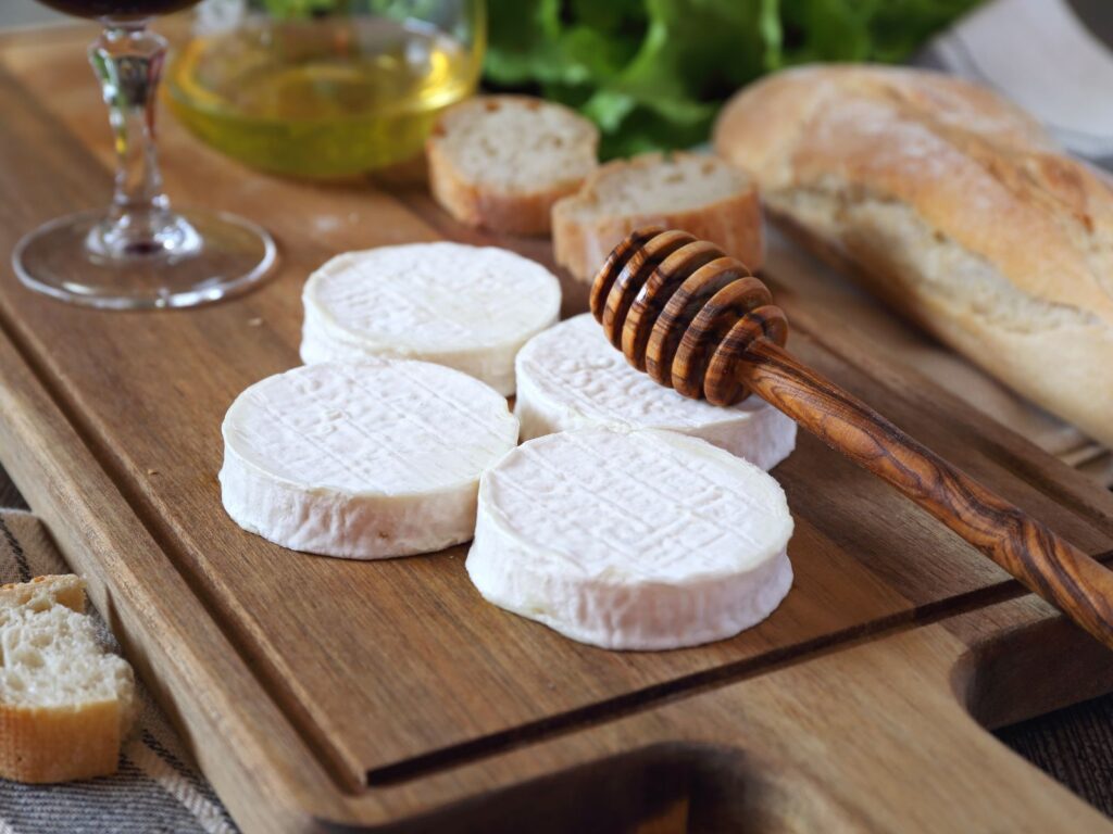Four small round white Rocamadour cheeses on a wooden board with honey dipper and baguette