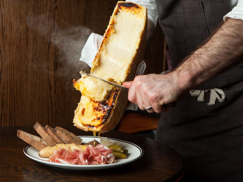 Waiter scraping melted Raclette cheese onto plate of cold meats