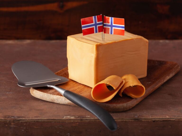 Block of Brunost topped with 2 Norwegian flags being thinly sliced